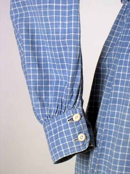 Klänning (bakförkläde) av blå-vit rutigt bomullstyg. Klänningen är rynkad runt om det släta oket. Oket är fodrat med ett vitrandigt bomullstyg. Manschetterna har sprund och knäpps med 2 trädda knappar. Framtill på höger sida finns ett hål för en ficka. I midjan finns en dragsko som knyts framtill på magen på insidan. Klänningen knäpps baktill med 7 hakar och sydda hyskor. Ett 11 cm brett skärp i samma tyg knytes runt midjan. Fläckar framtill samt ett hål.