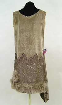 Klänning av ljusgrå silkesammet. På kjolen silverspets och längst ner grå plymer. I midjan en bukett tygblommor i rosa och blått. 1920-talet