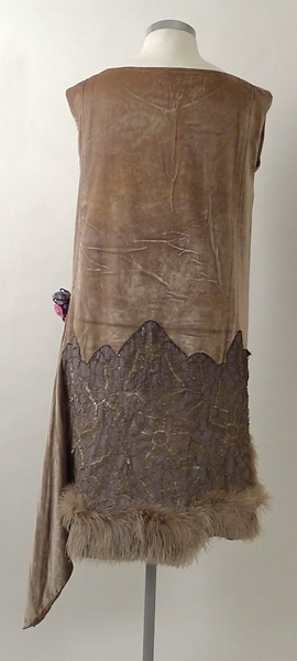 Klänning av ljusgrå silkesammet. På kjolen silverspets och längst ner grå plymer. I midjan en bukett tygblommor i rosa och blått. 1920-talet