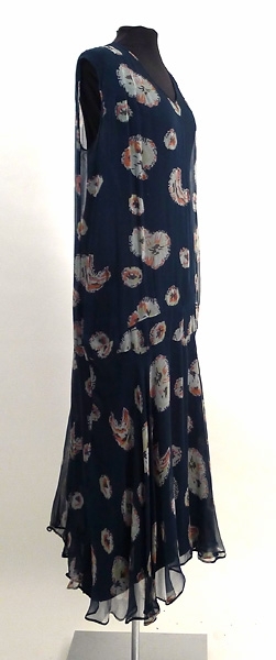 Enligt liggaren: Klänning med jacka. Blåblomig chiffon. Klockad kjol. Liv utan ärm med klockad volang. Tillhörande underklänning, svart crepe de chine. Tillhört Signe Påhlman