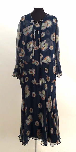 Enligt liggaren: Klänning med jacka. Blåblomig chiffon. Klockad kjol. Liv utan ärm med klockad volang. Tillhörande underklänning, svart crepe de chine. Tillhört Signe Påhlman