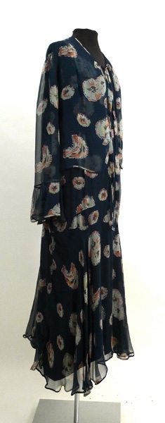 Enligt liggaren: Klänning med jacka. Blåblomig chiffon. Klockad kjol. Liv utan ärm med klockad volang. Tillhörande underklänning, svart crepe de chine. Tillhört Signe Påhlman