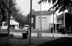 Från Lidköpingsutställningen sommaren 1946. 
Huset med Lidköpings stadsvapen och texten (Staden, Slätten, Vattnet) är Kyhlandersskolans norra sida. 
Fotografen står ungefär vid gatan, Esplanaden, och har skolans annex (ej synligt på fotot) på sin vänstra sida. 
Det lilla huset till vänster är en tillfällig paviljong. 
Damen till höger står på gångvägen mellan Esplanaden och Lasarettet. Ytterligare till höger ligger Stadsträdgården. 
Staden (rött), Slätten (gult), Vattnet (blått) var en slogan som en känd Lidköpingsprofil kommit på, Magnus Lidholm. Det röda är Lidköping färg. S:t Nicolaus är stadens skyddshelgon och brukar visas i röd beklädnad. (Jultomten). Gult för slättens gula säd och blått står för vattnet.
Plats: Kyhlanderskolans skolgård. Rätta namnet ska vara Västra Skolan, som den hette då.