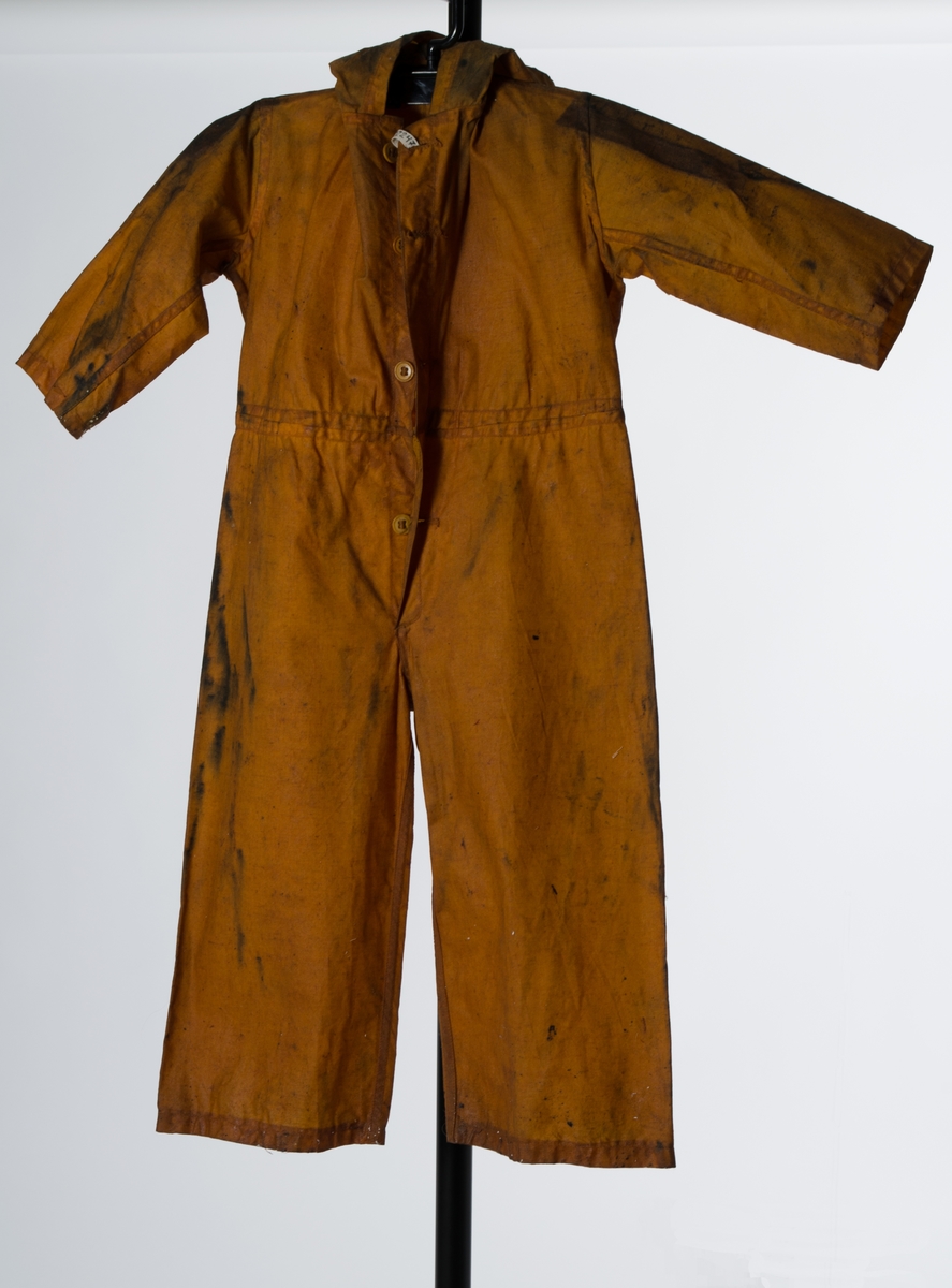 Okergul barnedress, sydd først, deretter dyppet i olje. Dressen har lange isydde ermer, kneppeåpning i front med fire knapper i ben (ant.). Løpegang i livet for stramming med snor. Påsydd hette. Doble sømmer.