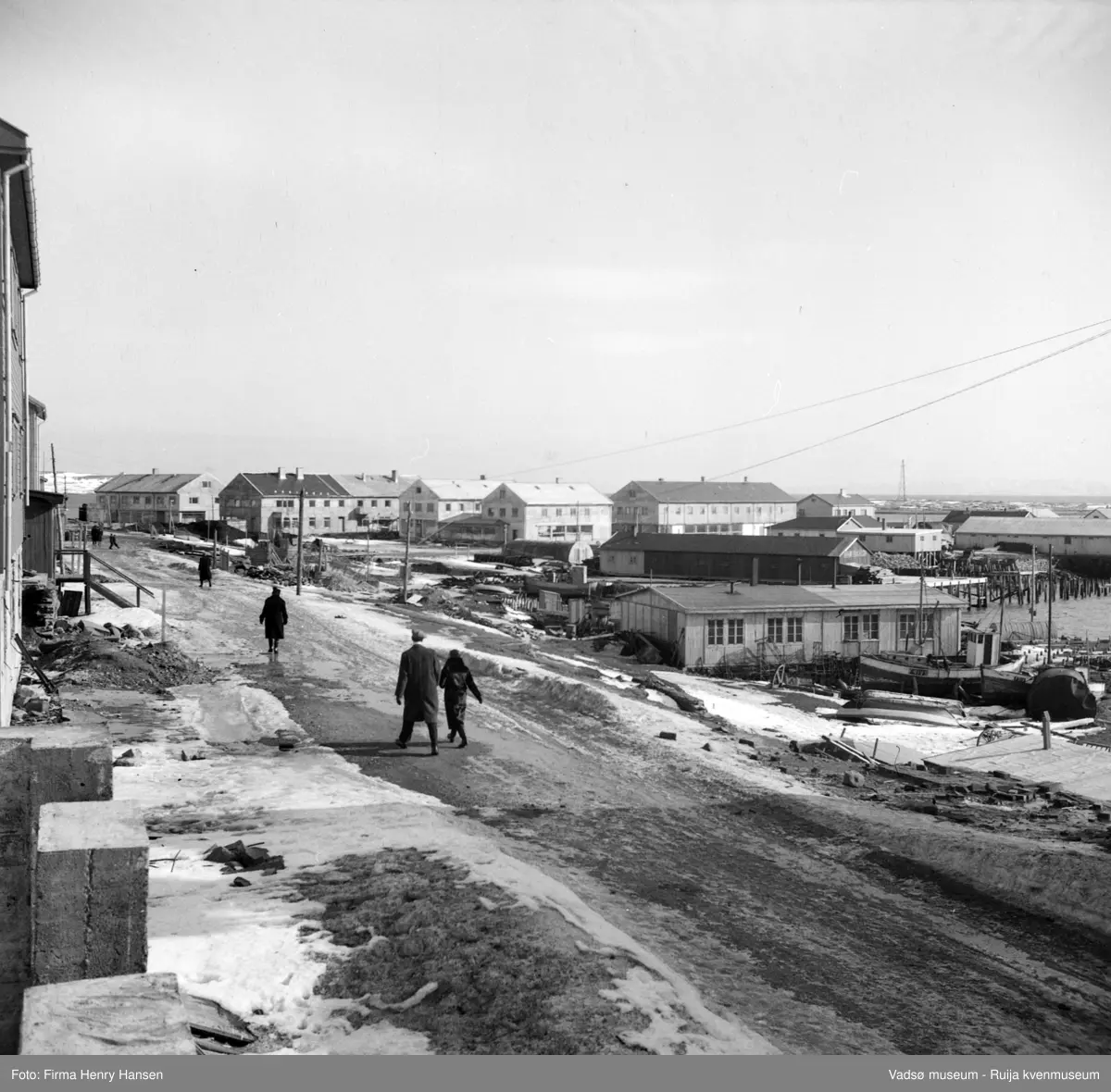 Vadsø sentrum fotografert fra nord, 1949. Det er vår og litt snø på bakken. Vadsø torg med forretningsbygg sør for torget: T. Mathisen, Leif Bjerck, baker Henriksen, Henry Hansen, Rolf Hansen og Esbensen. Det er brakker langs kaia, blant annet en Nissenbrakke. Luftskipsmasta på Vadsøya skimtes i bakgrunnen.