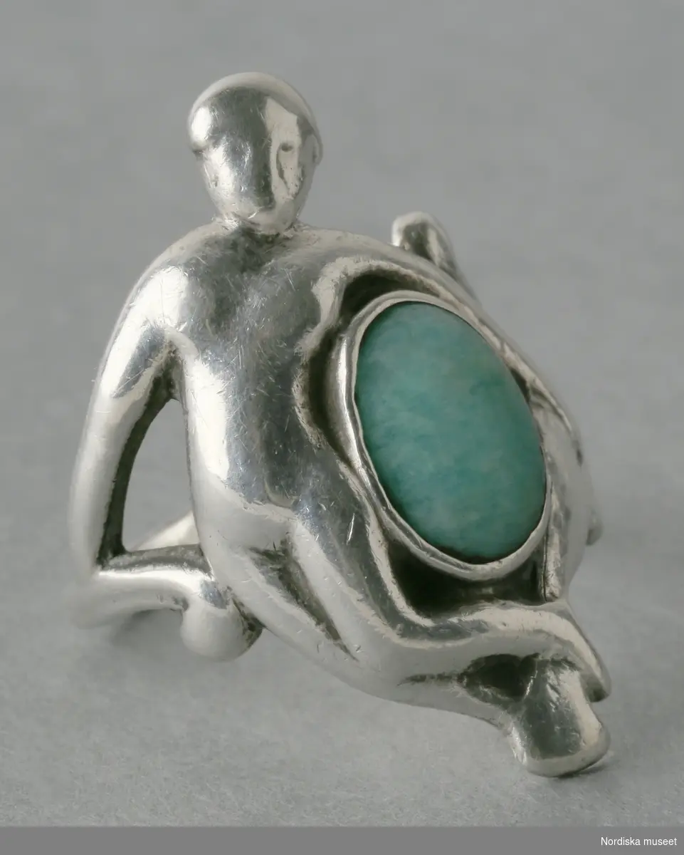 Ring (fingerring) av silver och grön sten. I jugendstil, föreställande kvinna och fisk. Stämplad med fyra silverstämplar: ett namnstämpel (IAR), svenskakontrollstämpeln (så kallad "kattfoten") med ett treklöverformade trekronorsvapnet, M7 (signifierar datumet) och ortsmärke ("L"). 
/Helena Lindroth & Petrine Knight 2014-01-22