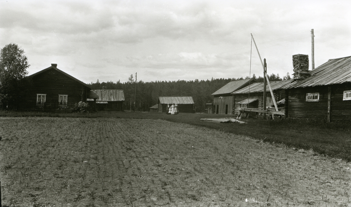 Gard, Østigarn (Østre Lutnes) m/gamle tømmerhus. Gammeldags brønn til høyre. Fire mennesker står midt utpå tunet (Tre kvinner og ei jente). Åker i forgrunnen.