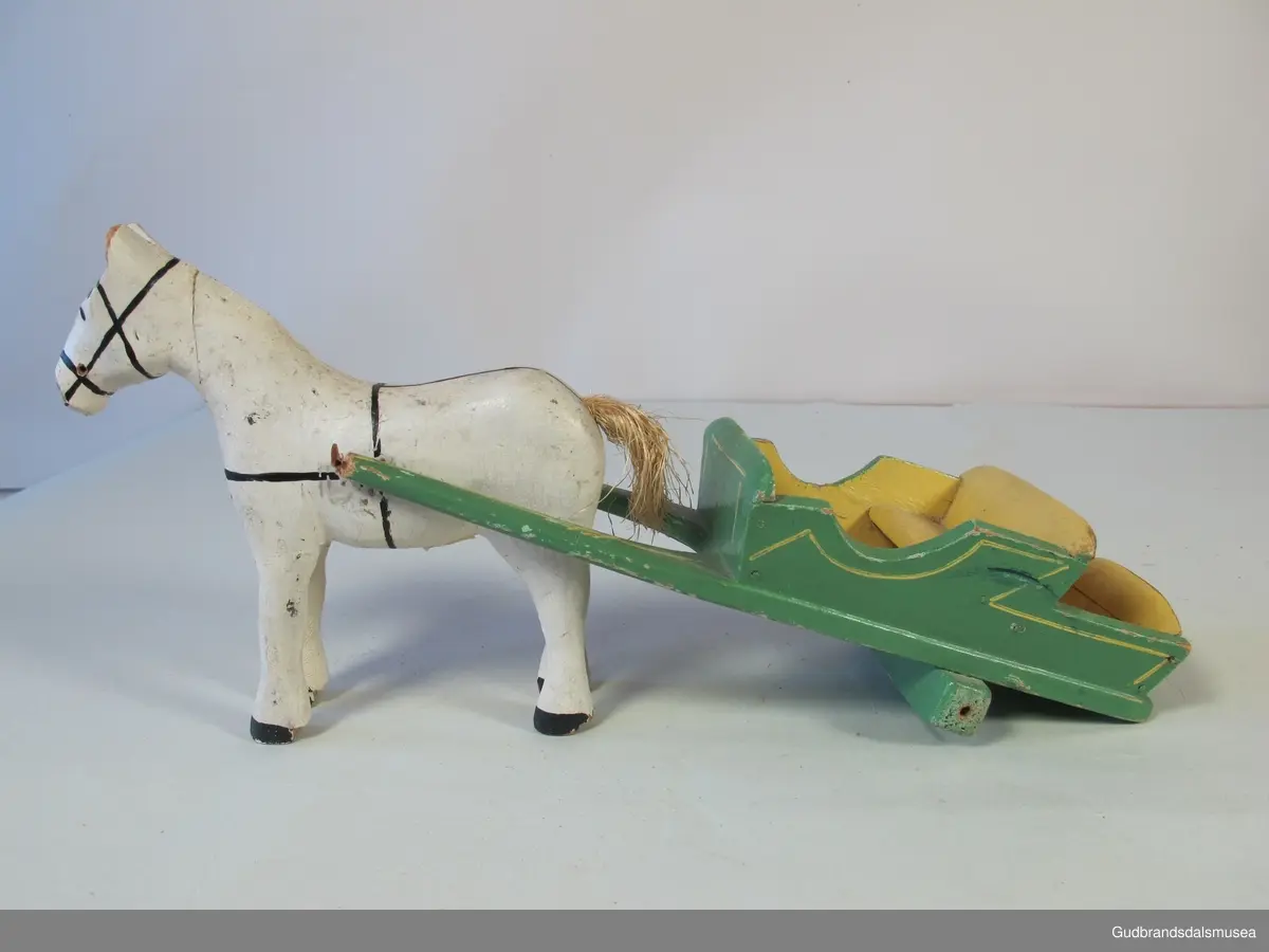 Trehest med kjerre. Begge deler malt. Hesten har påmalt sele og hale av stry. Skjekene er brekt der de har vært spikret til hesten, ig hjulene er vekk på kjerra/vogna.