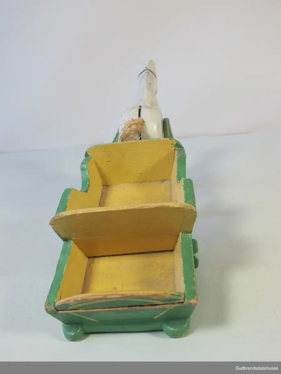 Trehest med kjerre. Begge deler malt. Hesten har påmalt sele og hale av stry. Skjekene er brekt der de har vært spikret til hesten, ig hjulene er vekk på kjerra/vogna.