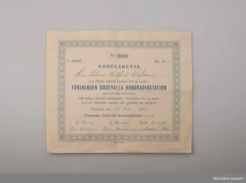 Andelsbevis i föreningen Uddevalla Rundradiostation U.P.A.
Vitt papper med blått påtryck inom dekorativ ram med texten:
N:r  1102
1 andel kr. 5:-
Andelsbevis 
Herr Edvin Walfrid Olofsson [handskrivet]
som tillfullo inbetalt insatsen för en andel i
FÖRENINGEN UDDEVALLA RUNDRADIOSTATION
utan personlig ansvarighet
tillförsäkras härmed delaktighet i föreningen för en andel
med de rättigheter stadgar och gällande lag medgiva.
Uddevalla den 31 dec. 1926
Föreningen Uddevalla Rundradiostation u. p. a. 
H. Elmér  G. Humble  Vald. Lindahl
Em. Sahlgren. Tycho Strömberg.