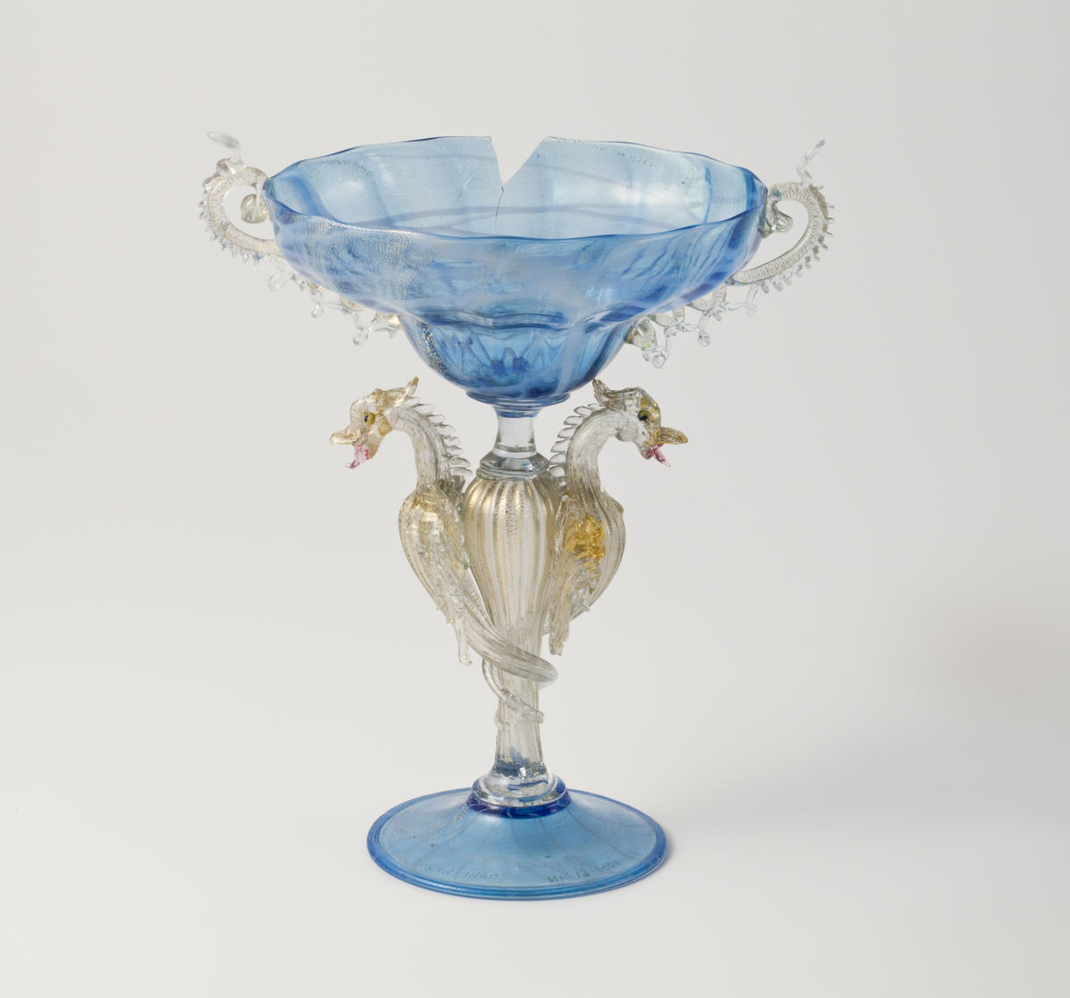 Prydglass med skålformet og vid kupa. Kupaen er blåfarget, mens hankene er utført i klart glass. Stetten er utformet som dragelignende vesener i klart glass med forgylt detaljering. Foten er sirkulær og blåfarget.