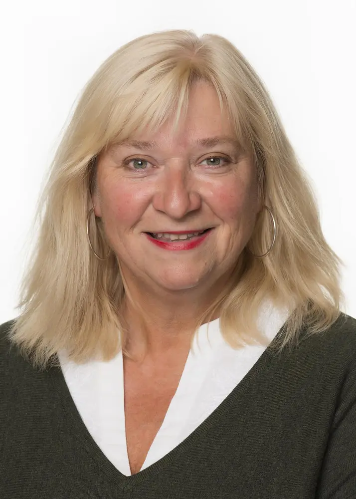 En kvinne med pannelugg, blondt hår til skuldrene, og sort og hvit genser smiler