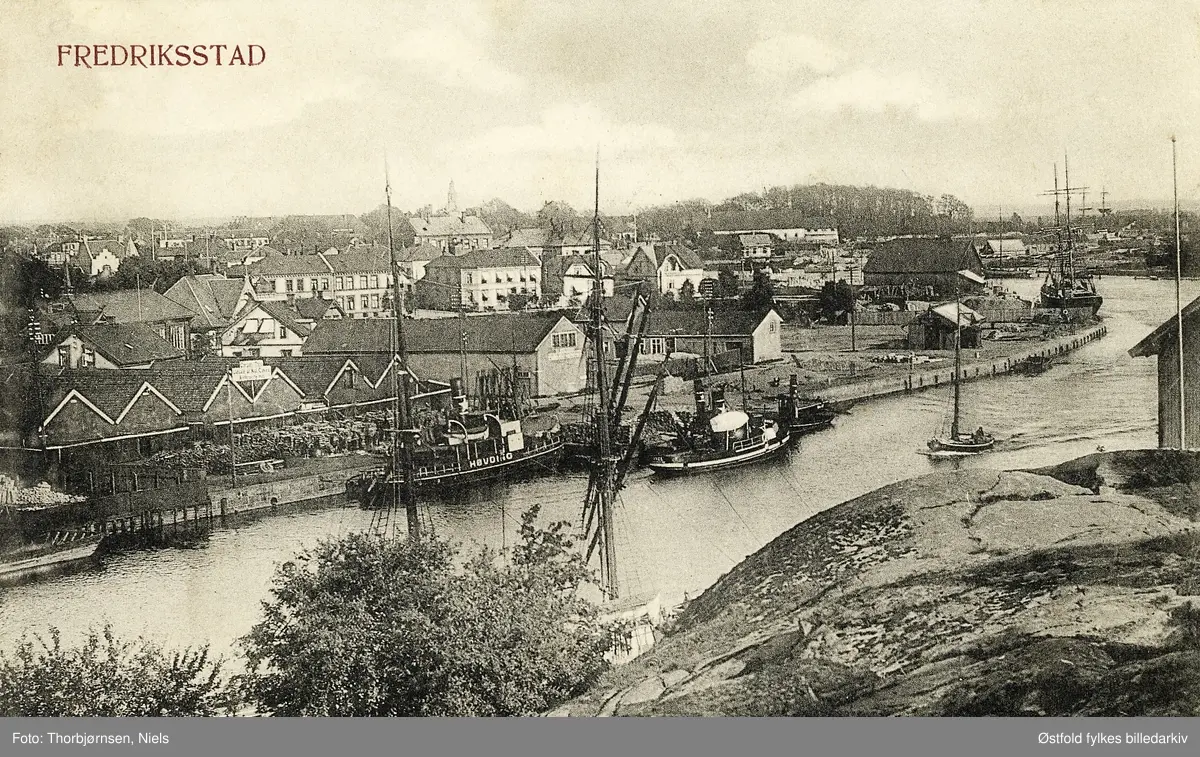 Vestsiden i Fredrikstad, prospektkort med uleselig poststempel.
Bildet er tatt fra Kråkerøy og viser Nygaardsbryggen med Cicignon og Gamlebyen bak.
Det vi ser til venstre langs elva er kullbodene til Carsten Thiis, hvor det ble solgt kull og koks. Vi ser Hiorthgården - eller Juelegården som den senere ble kalt etter at Julius C Juel overtok eiendommen i 1892.  Den ble revet i 1952 for å gi plass til Sentralsykehuset. (Kilde: Cicignon - fra festningsverk til villastrøk -red Svein Skahjem)
Slepebåt/taubåt MS Høvding ved kai. Båten ble bygget i Fredrikstad i 1893 som slepebåt, men på 1980-tallet ble ombygd til restaurantbåt og havnet i Egersund. Deretter havnet den som restaurantbåt / veteranbåt  i Flekkefjord.

Fra Norolf ellefsen 2017: 
Historien om "Høvding"kan ikke stemme,i følge Knut W Engebretsen som har skrevet om slepebåter i ælva,tilhører slepebåtene her C.A.Borgersen han eide en "Høvding" mellom 1905-17 den var bygd i Bergen i 1874 kom tilbake i til ælva i 1927 under navnet "Høvdingen",skrota her i 2014,eier Paulsen,Greåker I følge svensk register var Fmv's Høvding tømmersleper i Sverige,og var her en tur iår 2000 som "Fredrikshald", da norsk. Av dette følger at museet bruker feil "Høvding" i et bilde der den sleper barken "Fruen".


Fotografen står ytterst på Bjølstadfjellet, der det går ned i ''dær'n''  Dahl på Kråkerøy, her lå dengang Skribsreder Niels Thorbjørnsen sitt nedriggningsverksted av seilskuter, de ble rigget ned og bygget om til lektere, som igjen ble brukt til å frakte, takstein og muresten fra de mange teglverk langs elva, de ble også brukt til å frakte gatesten, sand, salt og andre ting som fyllte meget. Thorbjørnsen videresolgte så ankerkjetting, lanterner og tauverk fra de nedriggede skip til gjenbruk på andre skip.
På venstre side, den lange rekka med spisse hus, var lagerbygningene til Brukseier Carsten Thiis, som solgte byggematerialer som plank, knubb og kledning, men også ved Efter Thiis kom Fredrikstad Kullforretning (senere Nielsen & Sønner) Den litt større brakka lenger oppe ved seilskibet var byens Saltlager, det lå der faktisk frem til 1934 hvor det ble revet ned. Inn til venstre bak Thiis lagret kan vi se tårnet på Turnhallen, bak den skimter vi gavlen på J. N. Jacobsen's Lykkeberg. (Aage Dedekam forteller 2016)