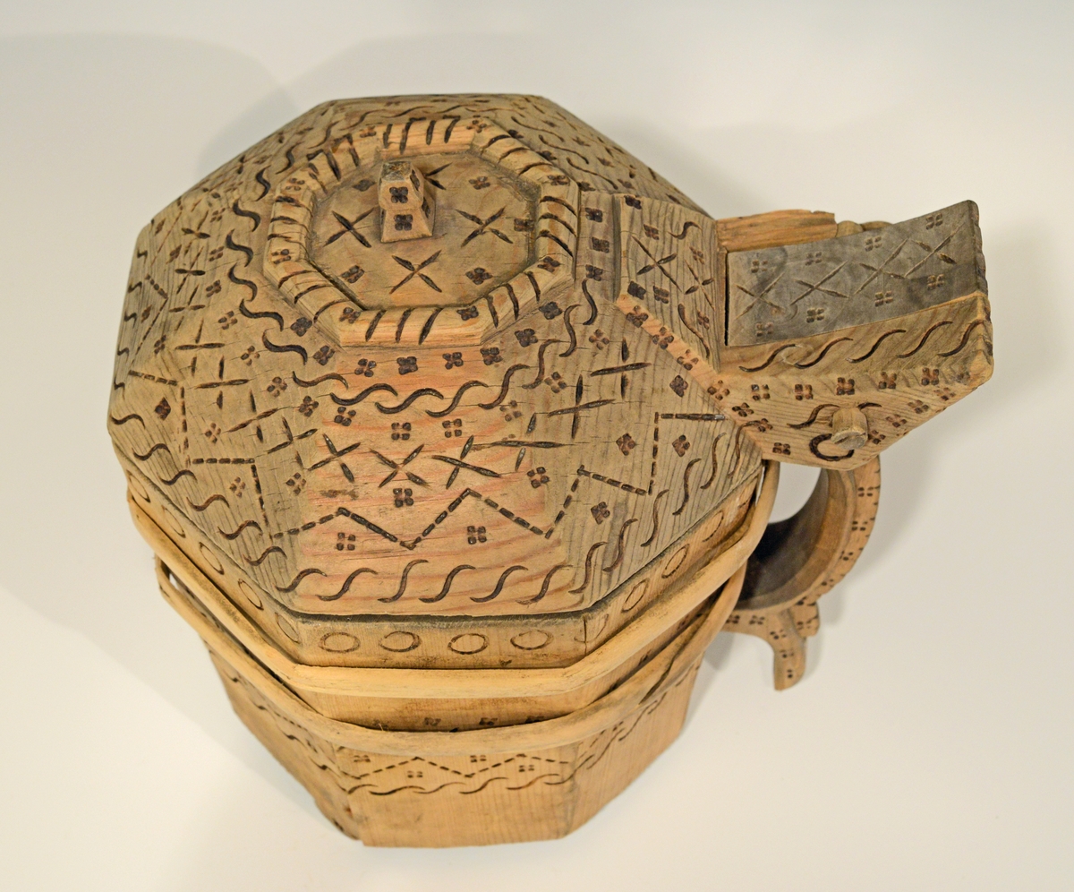 Fra katalog: Ottekantet smørkande og stav med hank og laak og tre smaa føtter, utvendig svimerket og en slynget ring oppe paa laaket, indvendig
rik rosette. Under staar brændemerket: A OSS 1823,