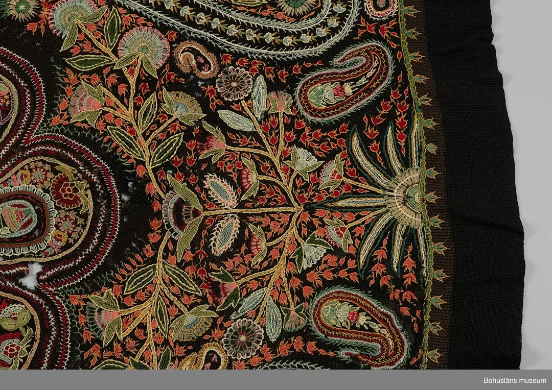 Överkast i mörk stickad (rätstickning) och virkad botten i ullgarn. Rikt broderad yta i kulörta färger (ullgarn färgat med anilinfärger).