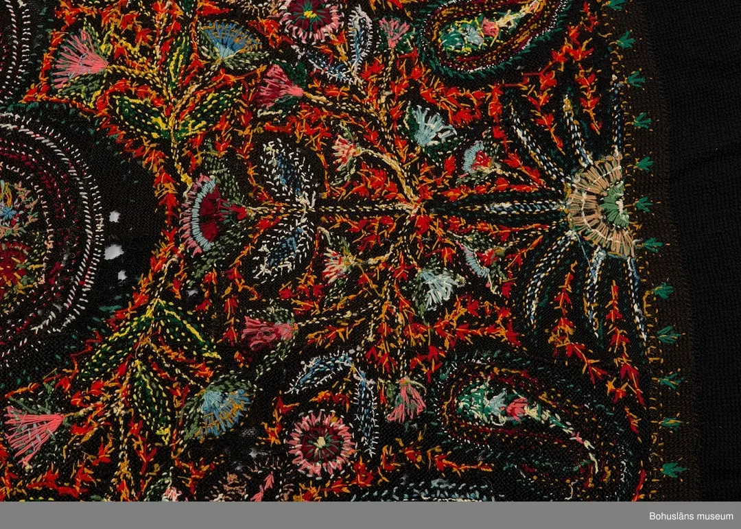 Överkast i mörk stickad (rätstickning) och virkad botten i ullgarn. Rikt broderad yta i kulörta färger (ullgarn färgat med anilinfärger).