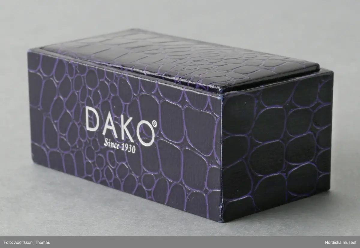 Etui av trä eller papp (stommen syns ej) klätt med lila skinnimiterande papper. På framsidan tryck "DAKO © / Since 1930" i silverfärg. Inuti klädd med grå syntettyg. Löst lock i samma material och utförande som etuiet. På ena långsidan en litet lila band av syntettyg (för öppnande) och på insidan av locket två öglor av resår (för att fästa manschettknapparna på). 
/Leif Wallin 2015-08-12