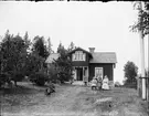 Man ligger i gräset, barn och kvinnor sitter på bänk framför Messings hus, Rovön, Uppland