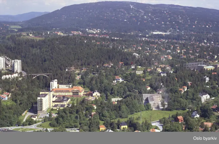 Radiumhospitalet til venstre, Ullern videregående skole til høyre.I venstre billedkant blokkene i Silurveien og trikkebroen over Merradalen, Ullernchausseen i front, Montebello og Husebyskogen i bakgrunnen. (Flyfoto)