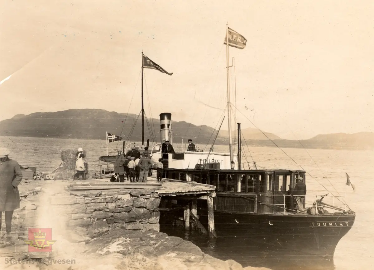 "Tourist ved Kvalvaag paa Frei 25.05.1926". I følge merking på baksiden av bildet. 
Se flere bilder fra befaringen: NVM 00-F-35784, NVM 00-F-35786, NVM 00-F-35787, NVM 00-F-35789 og NVM 00-F-35790, som alle er datert 25.05.1926 og registrert hver for seg.  

27.09.2018: "Tourist ved Kvalaag paa Frei" viser D/S "Tourist" fra Møre Fylkes Ruteselskap (MFR) ved Kvalvåg kai i daværende Frei kommune (nå Kristiansund kommune) med Kvisvika i daværende Straumsnes kommune (nå Tingvoll kommune) i bakgrunnen". 
Opplysninger fra Jon N. Eikrem, Stiftelsen Nordmøre museum, avd. Kristiansund.

27.09.2018: "Godt mulig at dette er en befaring i anledning vegen mellom Gyl (i Tingvoll kommune) og Treekrem (i daværende Straumsnes kommune) – gjennom Gylhamrene – som ble åpnet i 1931 (og utført som nødsarbeid). Sammen med ferjesambandet Kristiansund-Kvisvik (senere Kvalvåg-Kvisvik) som ble åpnet i 1932 (midlertidig utført med dampskip 1931-1932) skapte dette sammenhengende vegforbindelse mellom Kristiansund og Oppdal (dagens RV70). Før 1931 var det f.eks. ikke vegforbindelse mellom Straumsnes kommune og Tingvoll kommune. Ferjesambandet Kvalvåg-Kvisvik ble nedlagt ved åpningen av Krifast (fastlandsforbindelsen til Kristiansund) i 1992.  
Vesle D/S «Tourist» er nok innleid spesielt for denne befaringen (trafikkerte vanligvis ruta Kristiansund-Batnfjordsøra). Dampskipsruta på Tingvollfjorden (bl.a. med anløp av Øydegard og Sandvika) ble nedlagt i 1957".
Opplysninger fra Jon N. Eikrem, Stiftelsen Nordmøre museum, avd. Kristiansund. 

24.09.2018: "Bildet viser dampskipskaia der det seinere ble bilferge Kvalvåg-Kvisvik, fram til 1992". Opplysninger fra Odd Williamsen. 
24.09.2018: "Bildet må være fra Kvalvåg på Frei ja, med Kvisvika i bakgrunnen. Stemmer ikke med Kvalvåg i Gjemnes (da skulle vi sett Øydegard, Kjervika, Eikrem, Tveekrem og Treekrem rett over fjorden, og Aspøya med Boksaspa til venstre, og det er det ikke.) Opplysninger fra Jon Eikrem.  

"Kvalvåg er ei bygd i Kristiansund kommune, Møre og Romsdsal, som ligger på sørsiden av Freiaøya, vendt mot Freifjorden. Stedet hadde tidligere fergeforbindelse på MRF's rute Kvalvåg - Kvisvik over Freifjorden. Frei er den største øya i Kristiansund. (Wikipedia).
