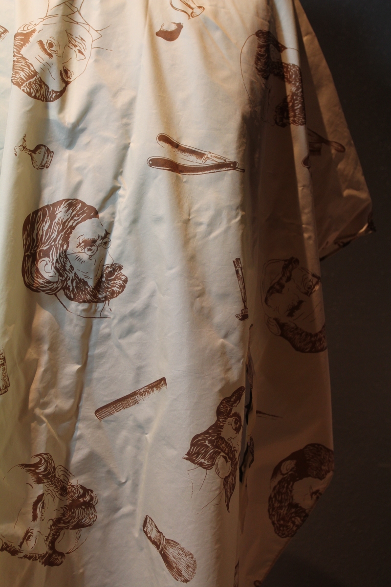 Form: avrundet, rektangulært stykke med halsringning, lukking bak. Stoffet har dekor som viser tegninger av barbersaker, børster og historiske menn med frisert hår, skjegg og/eller bart.