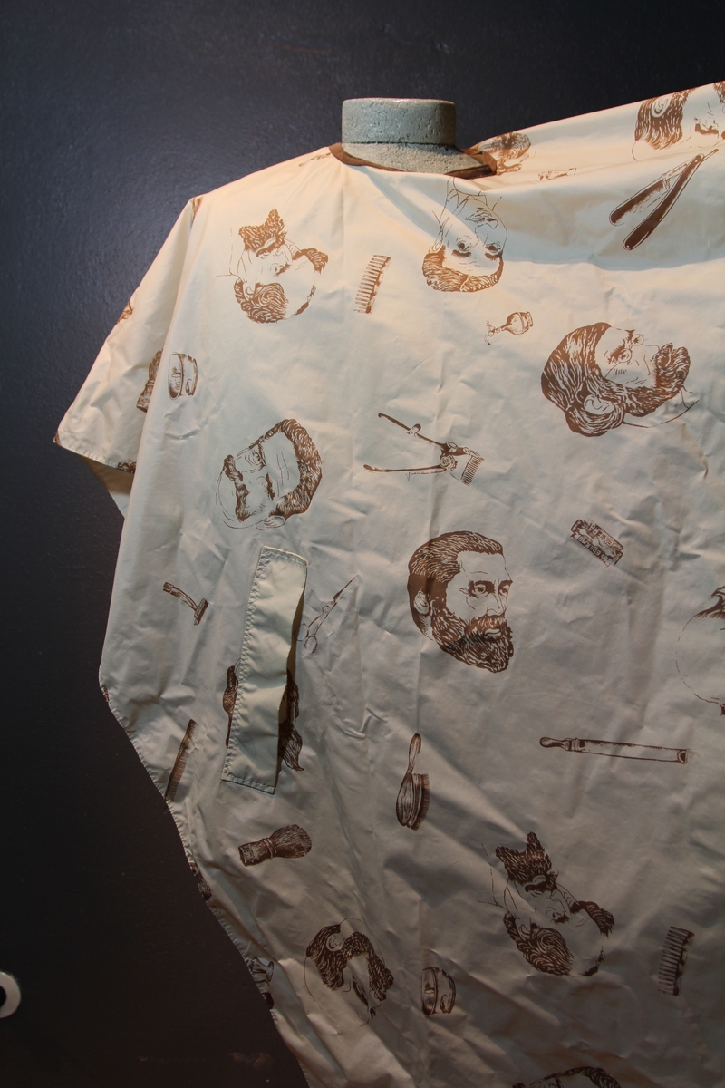 Form: avrundet, rektangulært stykke med halsringning, lukking bak. Stoffet har dekor som viser tegninger av barbersaker, børster og historiske menn med frisert hår, skjegg og/eller bart.