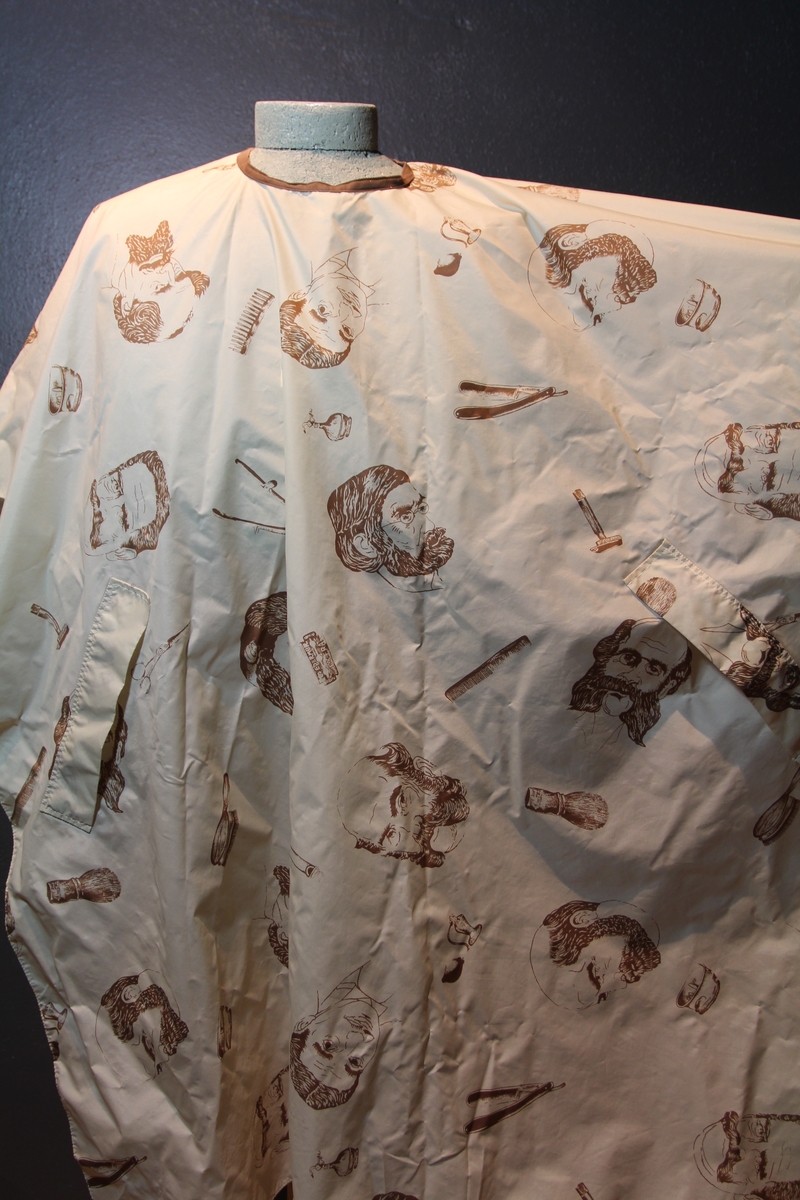 Form: avrundet, rektangulært stykke med halsringning, lukking bak. Stoffet har dekor som viser tegninger av barbersaker, børster og historiske menn med frisert hår, skjegg og/eller bart.