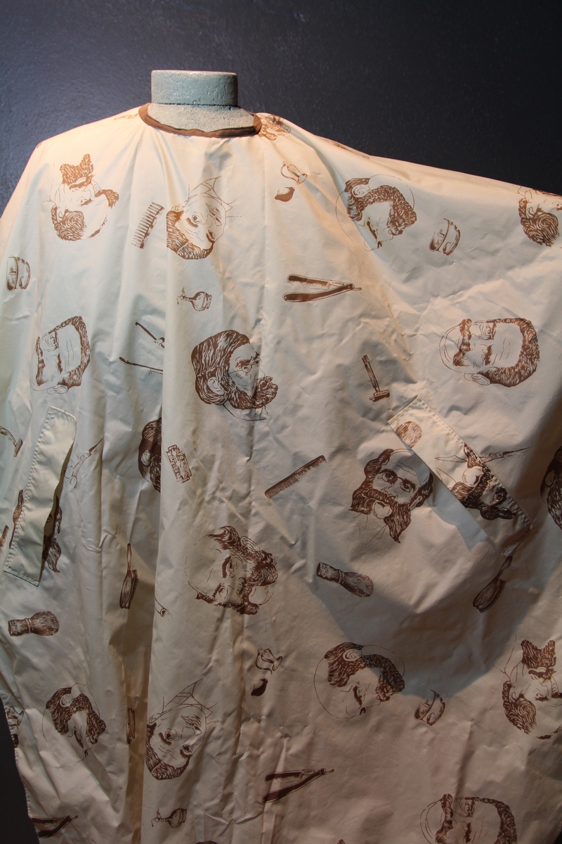 Form: avrundet, rektangulært stykke med halsringning, lukking bak. Stoffet har dekor som viser tegninger av barbersaker, børster og historiske menn med frisert hår, skjegg og/eller bart.