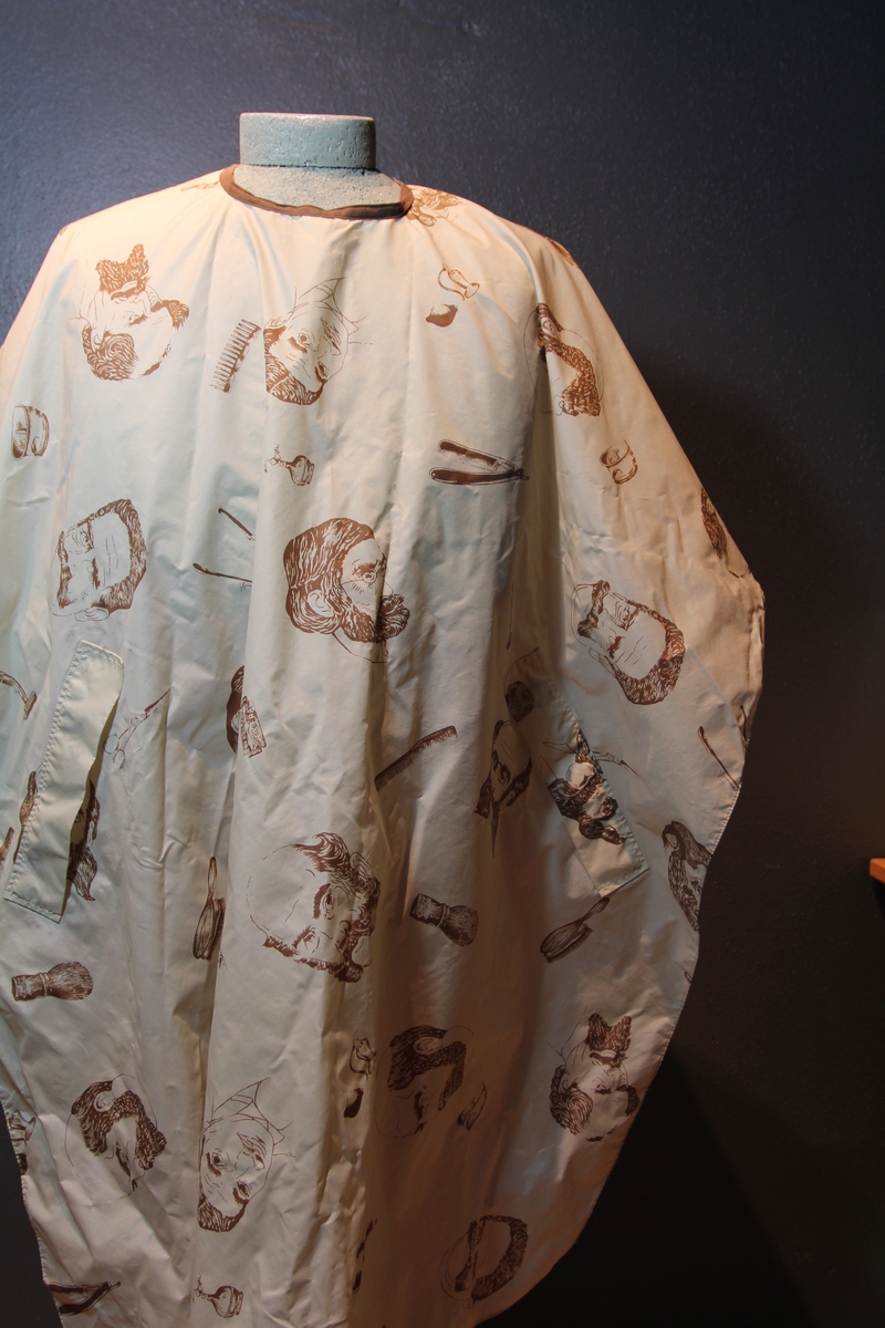 Form: avrundet, rektangulært stykke med halsringning, lukking bak. Stoffet har dekor som viser tegninger av barbersaker, børster og historiske menn med frisert hår, skjegg og/eller bart.