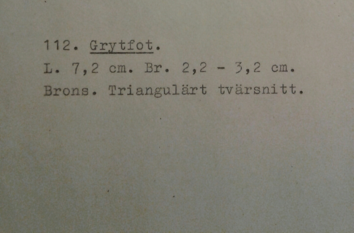 Grytfot av brons med triangulärt tvärsnitt. Breddad upptill mot grytans buk.