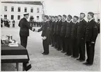 Uppställning av svenska marinsoldater på Sparregården i Karlkskrona. Amiral Tamm skakar handen med en marinsoldat och märker ut honom. På vänster sida ser man ett bord med fler utmärkelser som ska delas ut. I bakgrunden syns ett musikkår.