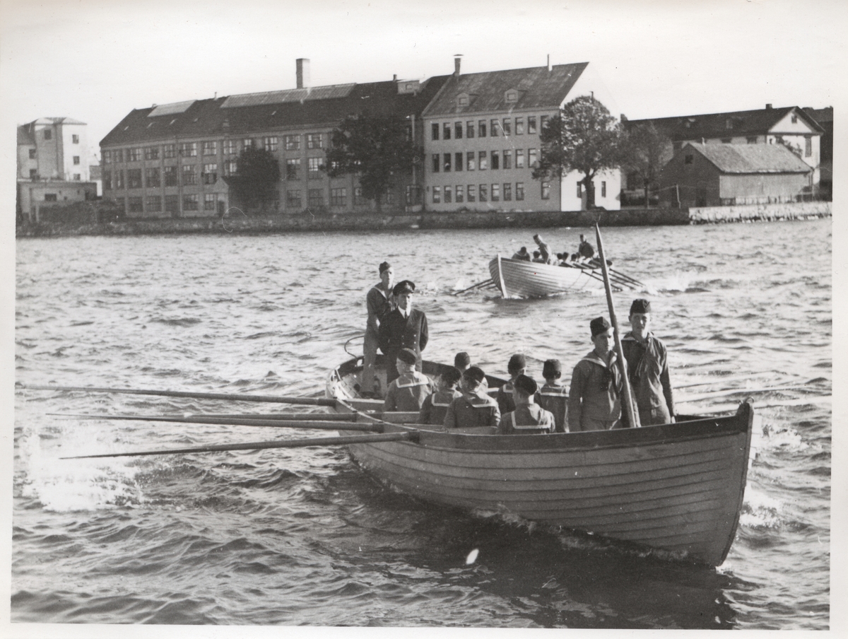 Centralt i fotografiets förgrund avbildas en klinkbyggd slup (åttahuggare) som ros. I fören står två unga män med en båtshake. Manskapet som ror sitter med ryggarna mot betraktaren och akterut står ett högre befäl (vilken tjänstgöringsgrad han har går dock inte att avgöra) och en ung man ur besättningen. Bakom denna båt, lite höger om bildens mitt, finns ytterligare en klinkbyggd åttahuggare som ros. Bildens bakgrund utgörs av Stumholmen; Beklädnadsförrådet (Kronobageriet), Beklädnadsverkstaden och Båtsmakasernen.