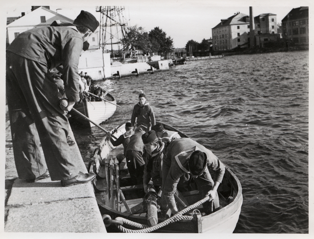 Fotografiet visar två slupar som har lagt till vid Kungsbron. I bildens omedelbara förgrund syns en av dem angjorda till kajen. Sex man ur besättningen är fortfarande ombord och iordningställer den. De får hjälp att lasta ur båten av kamrater som står uppe på kajen. Slupen bakom är delvis skymd av en av dessa, men det går att se att den matros som står i fören har båtshaken i beredskap. Den högra delen av bilden, samt det mesta av förgrunden, utgörs annars av vatten. I bakgrunden ses Matinrättningen, Beklädnadsförrådet (Kronobageriet) samt Beklädnadsverkstaden.