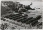 Uppställning av manskap i kolonner på stor plan under medaljutdelning. I bakgrunden går båtar och fartyg samt natur i form av skogsdunge. Bilden är tagen högt uppifrån.
