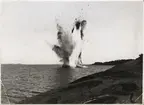 Fotot visar sprängning av fartyg. Explotionen orsakar ett stort moln av rök blandat med delar av båten. I förgrunden och bakgrunden syns klipphällar, hav och klar himmel.