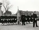 Militärer i uniform på stortorget i Karlskrona framför Tyska kyrkan. Sjöbefäl står i täten varav en håller upp svenska flaggan. Matroserna bär eldvapen och sjöbefälen värjor.