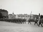 Militärer marchera på stortorget. I täten går flaggbärare som bär Sveriges flagga. I bakgrunden syns åskådare och bebyggelse.