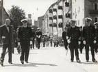 Sjöbefäl och civila promenerar nedför gata. I bakgrunden syns bebyggelse med bl.a Fredrikskyrkans torn.