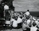 Folksamling sitter på gräsbeklädd sluttning och ser ut över havet som skymtar i höger bakgrund. Två män, varav den ena uniformsklädd, står vid en kamera på stativ. Bilden är tagen i samband med 
