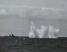 Under en övningsskjutning syns ett fartyg syns på avstånd från kameran intill en explosion i havet som kastar upp vattenmassor i luften. Bilden är tagen i samband med 