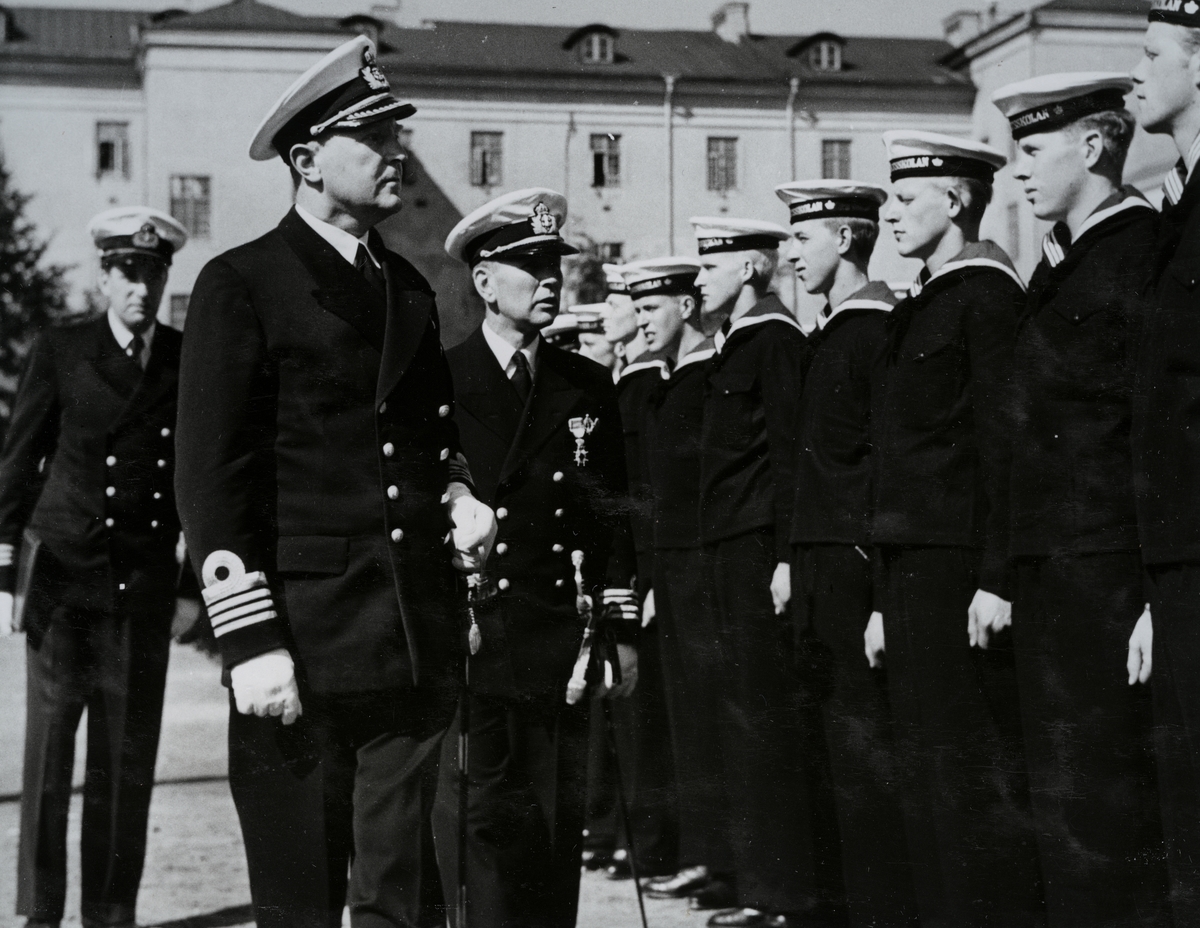 Uniformsklädda flottister står uppställda på rad inne på Sparres kaserngård. Framför dem går tre äldre uniformsklädda män med högre gradbeteckning, den främsta av dem är kommendör Bong.