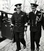 Konteramiral Johan Erik Samuelson promenerar tillsammans med en högre officerare. Båda är iklädda uniformer. Vid sidan om dem samt i bakgrunden syns två bilar längs gatan samt fler män i militära högtidsuniformer.