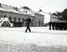 En grupp flottister och en kapten marscherar över Sparres kaserngård. I bakgrunden syns byggnaden för aula, gymnastiksal och utbildningsförråd. Framför byggnaden står en rad av högre militärer och på en flyttbar trappa står ytterligare en högre militär och gör honör.