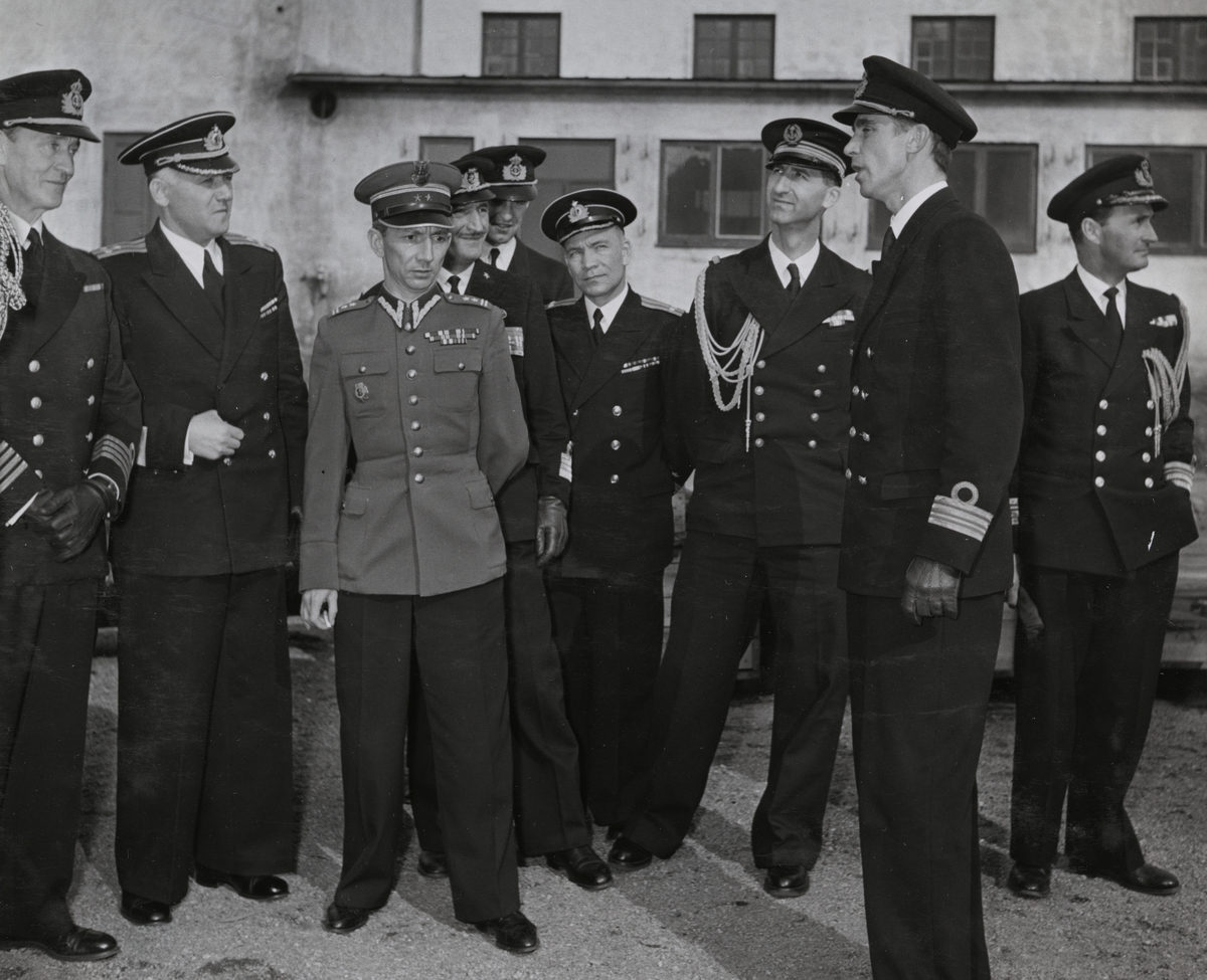 Kommendörkapten Lind af Hageby (till vänster) står tillsammans med en grupp höga militärer på en grusyta utanför en byggnad. Samtliga är klädda i uniform.