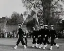 En grupp uniformsklädda flottister, den första med en svensk flagga,  marscherar över Stortorget i Karlskrona. I bakgrunden syns vattenborgen och en folksamling som har samlats för att titta på marschen.