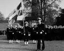 Sjömän står uppställda. En kapten går i förgrunden och bakom honom, i spetsen på de uppställda soldaterna står en fänrik hållandes en stor, tretungad svensk flagga. I bakgrunden syns vattenborgen på stortorget bakom ett träd.