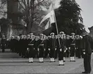 Uniformsklädda flottister står uppställda på Stortorget framför vattenborgen i Karlskrona. Fänriken längst fram håller i en svensk flagga.