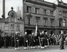 Uniformsklädda militärer från KA:2 står uppställda på Stortorget framför Karlskrona stadshotell. I bakgrunden framför hotellet står en folksamling bestående av barn och vuxna som tittar på.