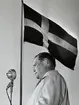 En civilklädd man står bakom en mikrofon framför en flaggstång med en hissad svensk flagga.