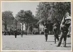 Soldater står på grusgården framför Jämjö Gamla Kyrkskola, som skymtar till höger i bild. Framför dem står ytterligare en hemvärnssoldat i uniform samt en präst. Bakom dem, med ryggarna mot Jämjö Kyrka som syns i bakgrunden, står en musikkår. Runtom står civila åskådare.